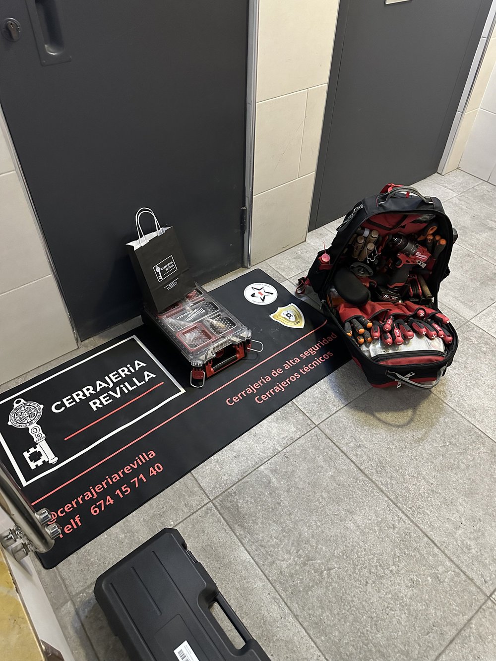 Herramientas de cerrajería en el suelo, incluyendo una mochila roja con herramientas y una bolsa negra junto a una puerta; alfombra negra con el texto "Cerrajería Revilla" en el piso.